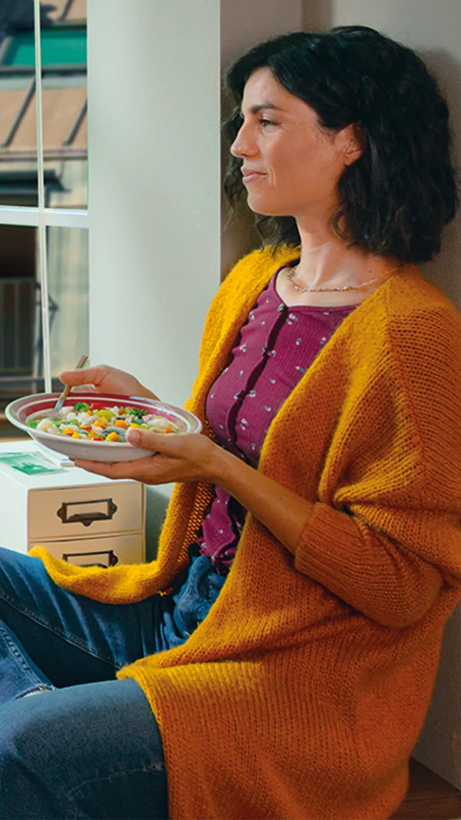 Donna seduta sul davanzale di una finestra, con un cardigan giallo e jeans, tiene in mano una ciotola di insalata mentre guarda fuori; accanto a lei una scrivania con libri, quaderni, una lampada e portapenne in un ambiente luminoso e accogliente.