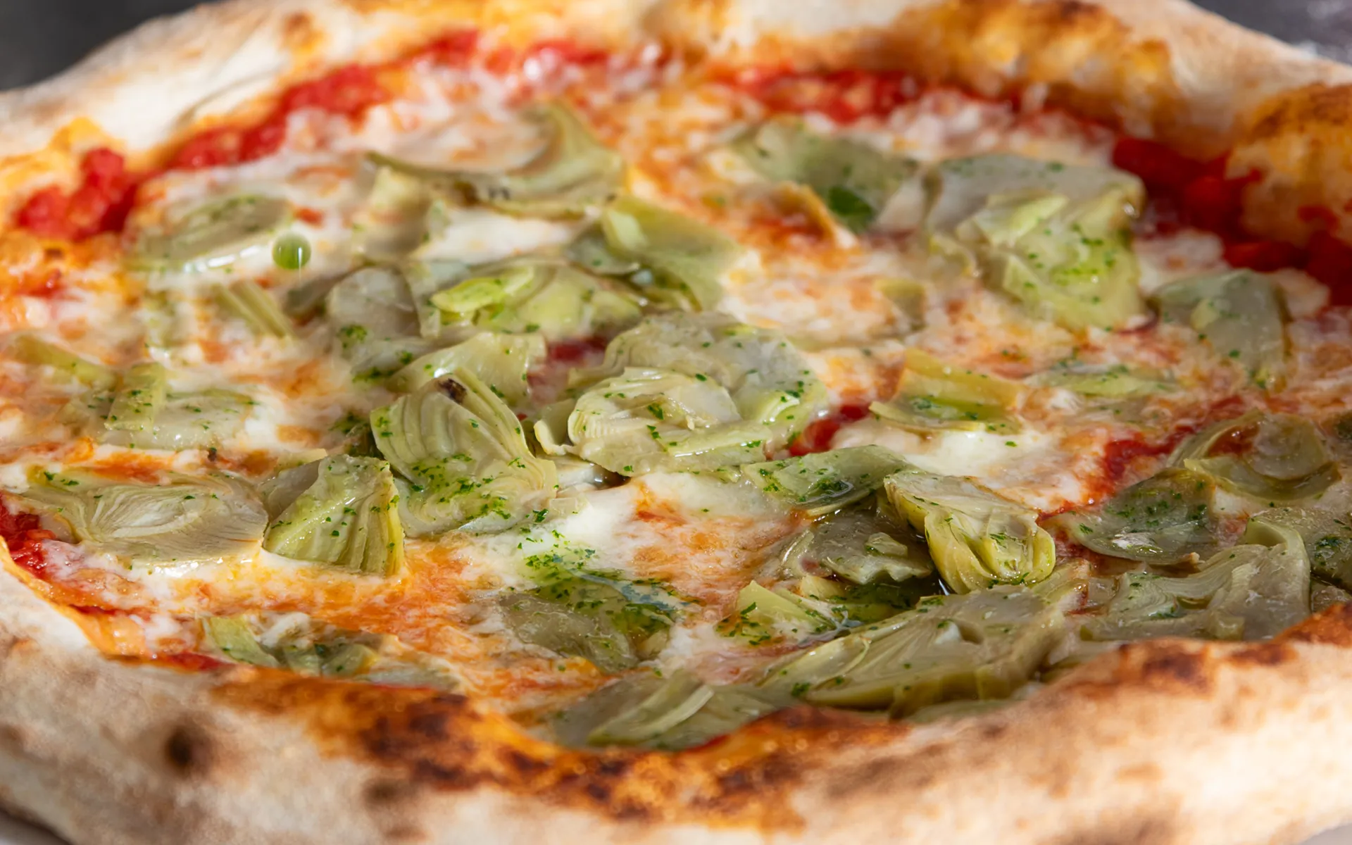 Pizza con pomodoro, mozzarella filante, carciofi a fette e prezzemolo, dal profumo fresco e mediterraneo.