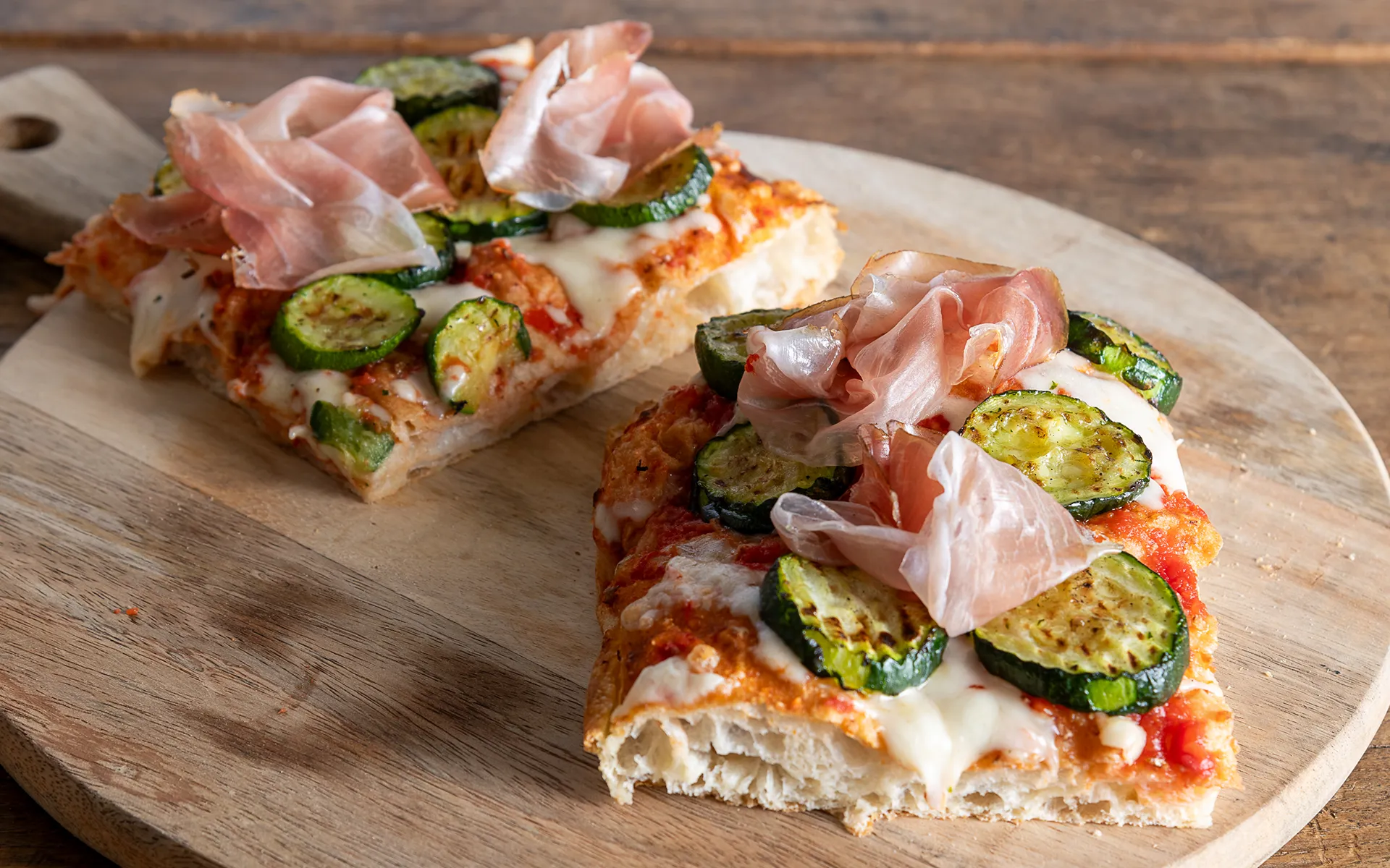 Pizza in teglia con pomodoro, mozzarella filante, zucchine grigliate e speck, dal gusto equilibrato e deciso.