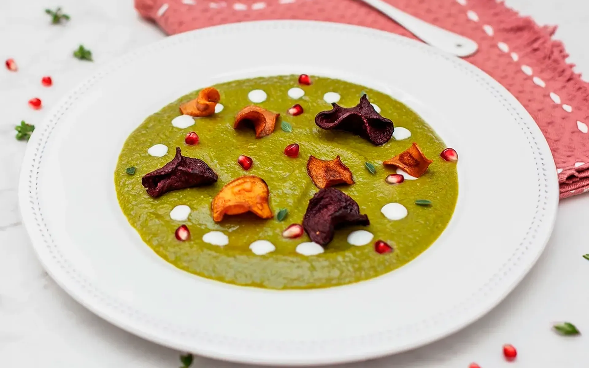 Passato di verdure con chips croccanti e melagrana, servito su piatto bianco.