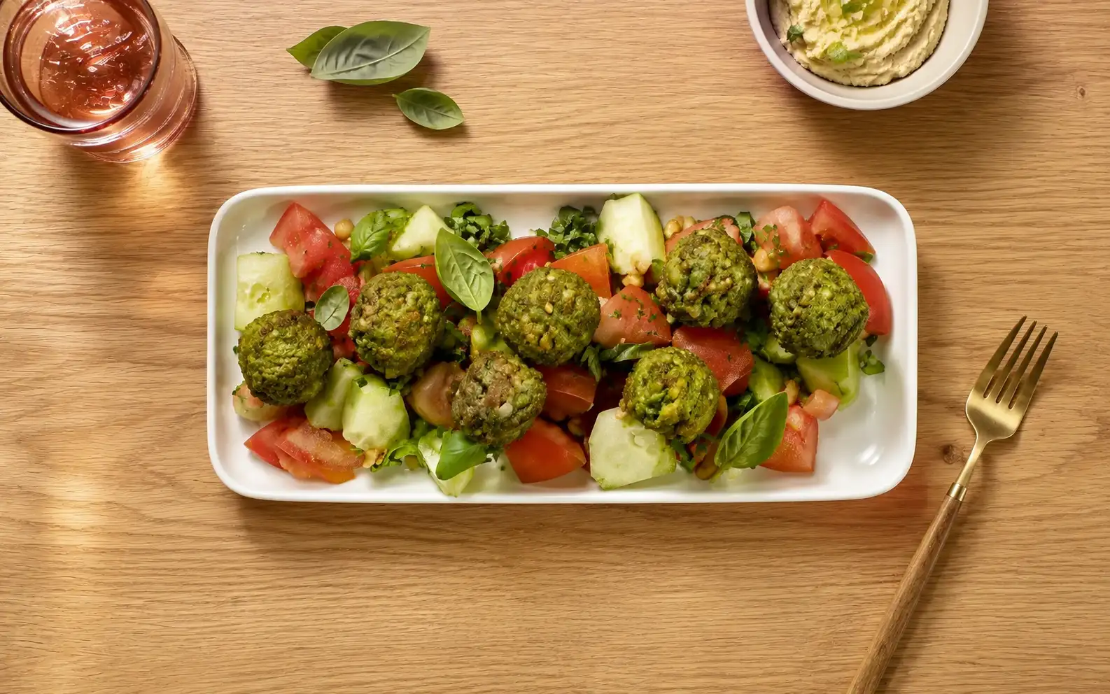 Insalata fresca con polpette vegetali agli spinaci, pomodori, cetrioli e basilico, servita su piatto bianco e fotografata dall'alto su tavolo in legno.