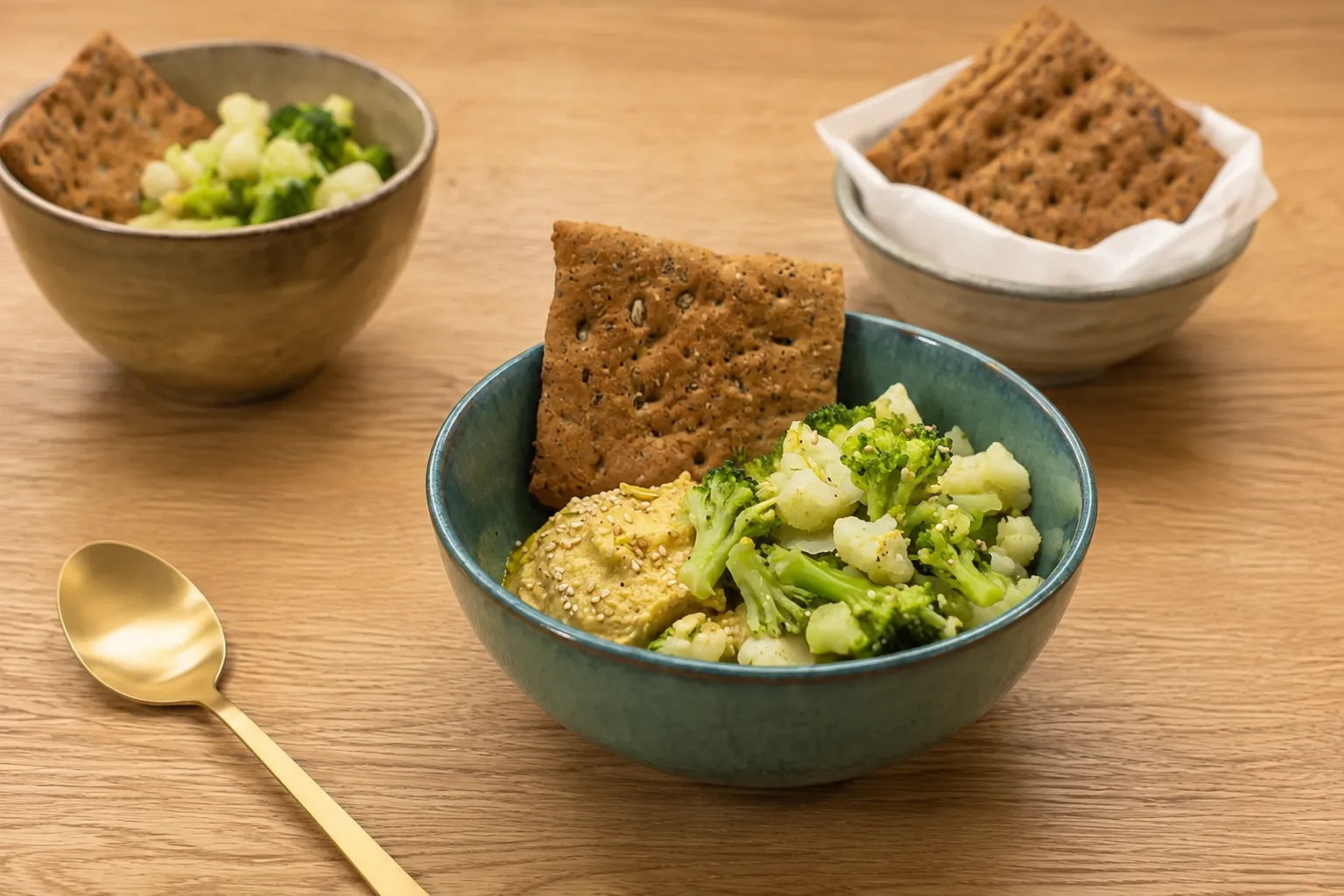 Ciotola di hummus con broccoli e cavolfiore, accompagnata da cracker integrali e fotografata su un tavolo in legno di rovere con atmosfera calda e naturale.