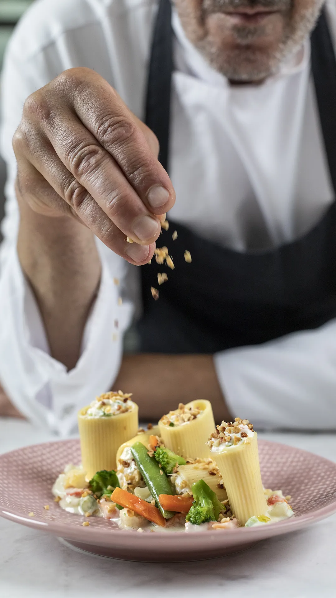 Chef che completa un piatto di paccheri ripieni con verdure e crema, aggiungendo granella croccante sopra la pasta.