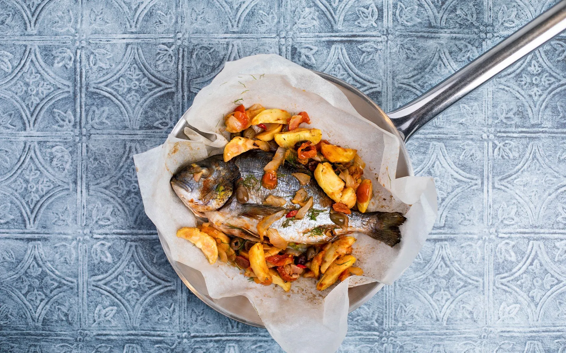 L'immagine mostra una padella vista dall'alto, contenente un orata al forno con patate., alla cacciatora con olive taggiasce e aneto