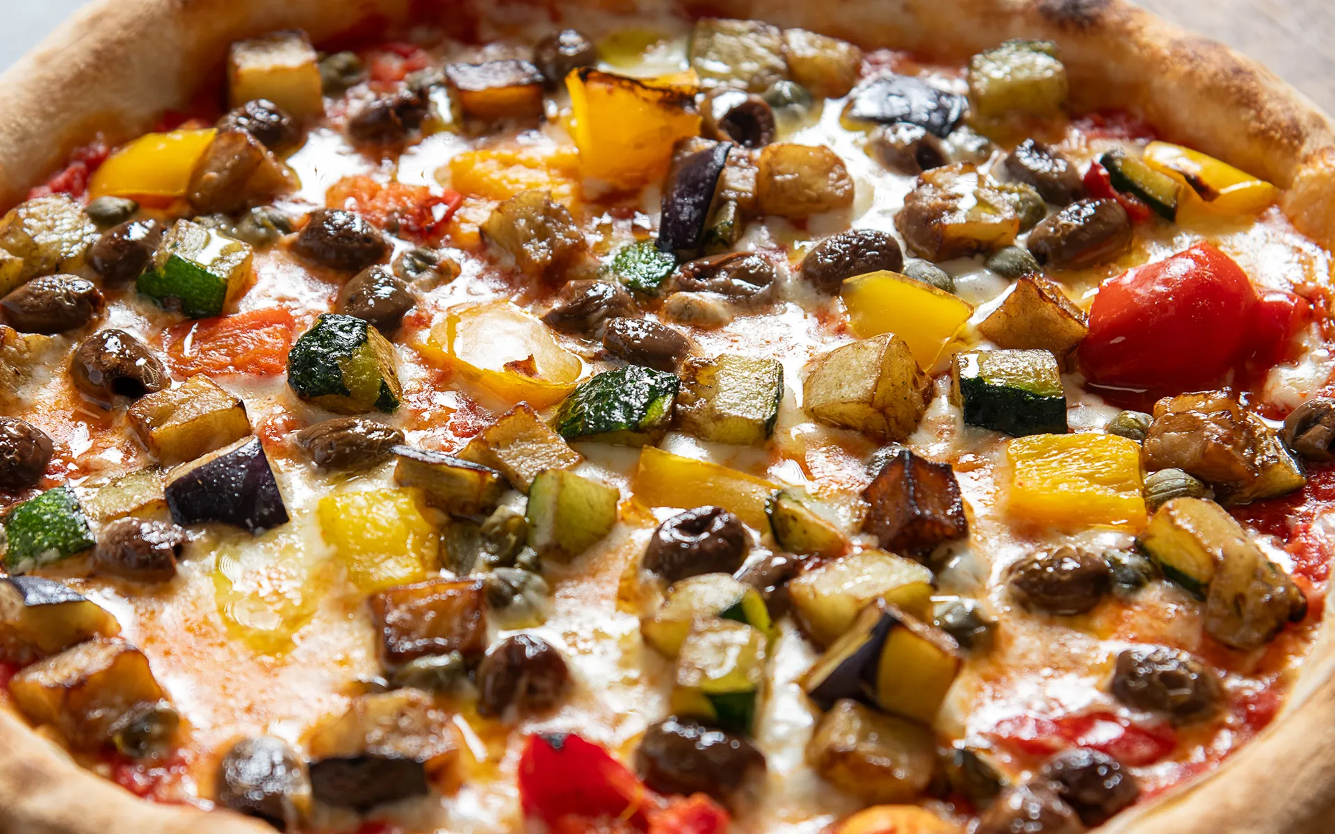 Pizza tonda con pomodoro, mozzarella filante e verdure alla mediterranea con capperi e olive taggiasche, dal gusto intenso e aromatico.