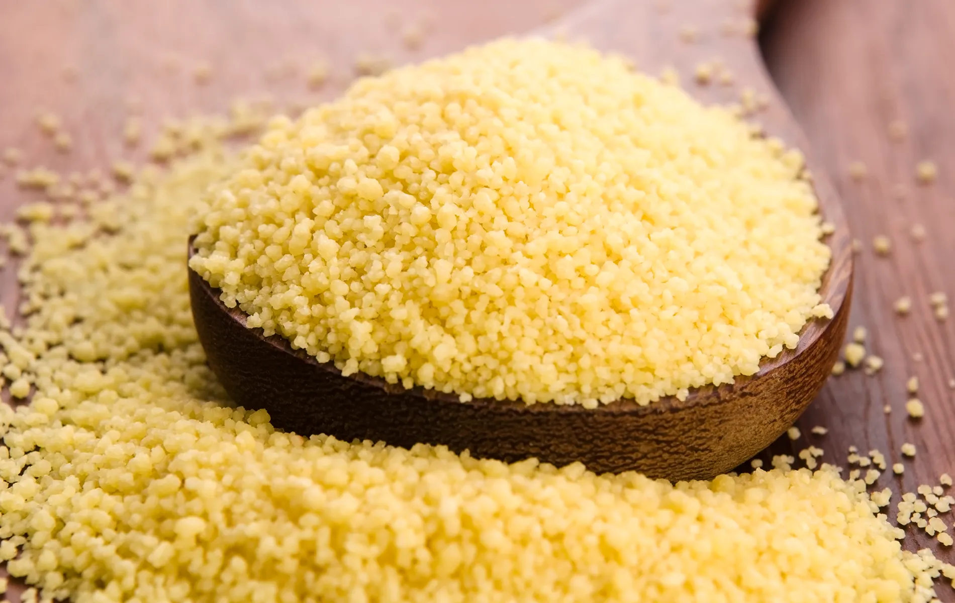 Cucchiaio di legno colmo di cous cous crudo su tavolo in legno, con piccoli granelli sparsi attorno.