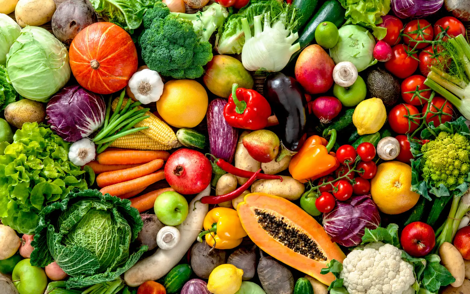 Assortimento di verdura e frutta fresca disposto su un tavolo in legno visto dall'alto: broccoli, cavolfiore, pomodori, peperoni, melanzane, carote, cavoli, finocchi, zucchine, patate, zucca, funghi, agrumi, mele e altri ortaggi colorati.