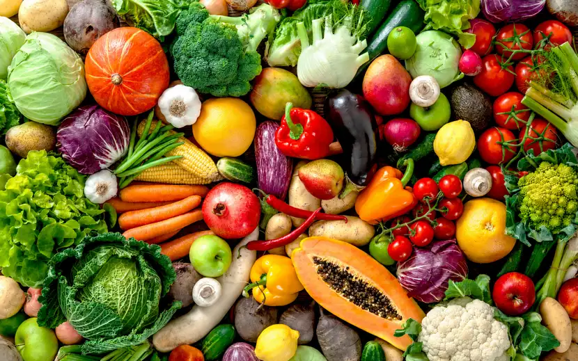 Assortimento di verdura e frutta fresca disposto su un tavolo in legno visto dall'alto: broccoli, cavolfiore, pomodori, peperoni, melanzane, carote, cavoli, finocchi, zucchine, patate, zucca, funghi, agrumi, mele e altri ortaggi colorati.